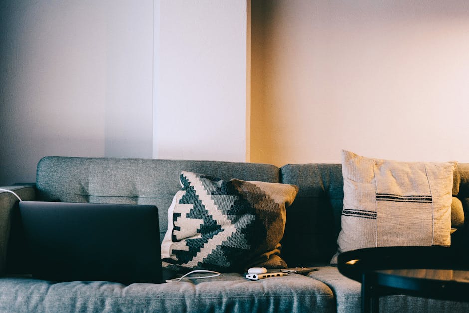 A cozy living room setup featuring a sofa with pillows and a laptop, perfect for relaxation and remote work.